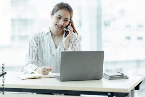 Cheerful young Asian woman smiling and looking away while having smartphone conversation in office