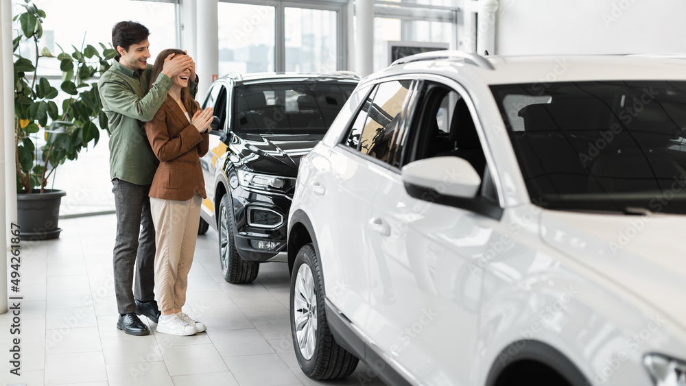 Handsome young man covering his wife's eyes, making surprise gift, buying new car at automobile dealership store Handsome young man covering his wife's eyes, making surprise gift, buying new car at automobile dealership store