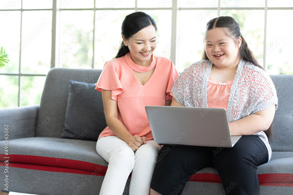 mother using laptop computer and a girl with down syndrome or her daughter, smiling and enjoying on sofa mother using laptop computer and a girl with down syndrome or her daughter, smiling and enjoying on sofa
