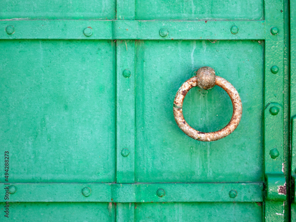 Green metal gate. The door handle in the form of a ring is rusted. Close-up. Rough iron doors painted green. Green metal gate. The door handle in the form of a ring is rusted. Close-up. Rough iron doors painted green.