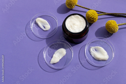 Petri dishes with white cream smears and cream in dark glass jar on violet background with yellow flowers of craspedia.