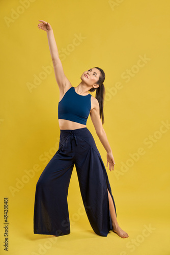 Front view of young woman standing barefoot, raising hand, looking up. Brunette girl with pony tail stretching, practicng yoga pose. Isolated on yellow studio background.  photo