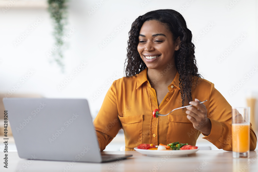 Cheerful african american woman having snack in front of laptop