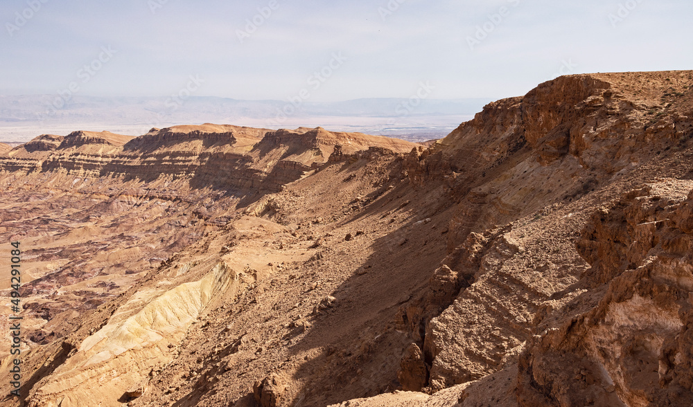 section of the south rim of the Makhtesh Katan Small Crater showing cliffs and colorful sandstone with the Jordan Rift Valley in the background