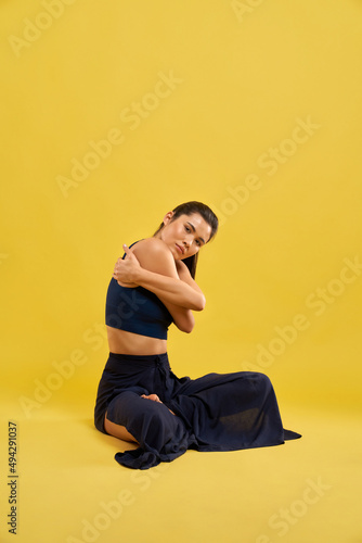 Front view of sporty girl sitting with crossed legs, holding by hands, hugging, looking at camera. Brunette wearing sport suit, doing yoga pose. Isolated on yellow background. © serhiibobyk