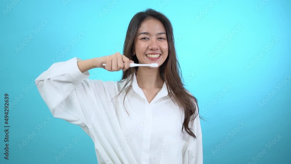 Front view half-body image of a happy beautiful Asian woman standing holding a toothbrush and demonstrate the gesture of brushing her teeth while smiling broadly at camera isolated on blue background. Front view half-body image of a happy beautiful Asian woman standing holding a toothbrush and demonstrate the gesture of brushing her teeth while smiling broadly at camera isolated on blue background.