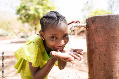 Smiling young African girl at a faucet, letting water flow on her hands, while drinking. photo