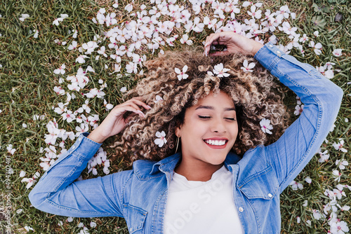 Murais de parede top view of happy hispanic woman with afro hair lying on grass among pink blossom flowers