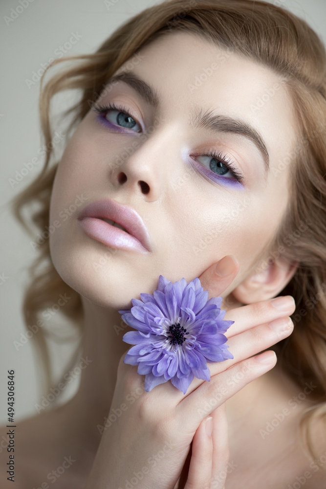 Beautiful young woman's face with perfect skin and makeup holds beautiful blue spring flower close to her face. Spring seasonal concept for SPA, skincare and wellness. Close-up, selective focus Beautiful young woman's face with perfect skin and makeup holds beautiful blue spring flower close to her face. Spring seasonal concept for SPA, skincare and wellness. Close-up, selective focus