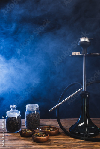 Hookah near dry tea in bowls on wooden surface on black background with blue smoke.