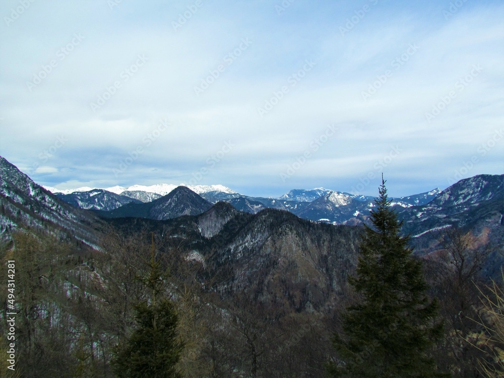 View of forest covered hill in Karavake mountains from the top of Javorjev Vrh in Gorenjska, Slovenia in winter with mountain peaks in the back covered in snow
