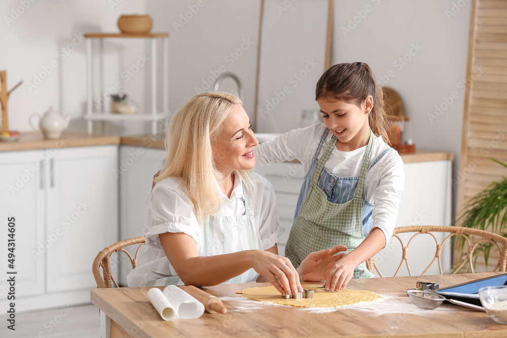 Mature woman and her little granddaughter cutting dough for cookies at table in kitchen