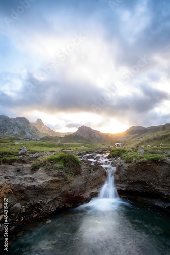 a meadow on top of a mountain  surrounded by rocky peaks  with a small river ending in a small waterfall in the foreground  cows grazing  mountain sunset on a cloudy day  vertical