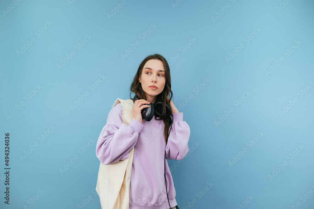Attractive woman in casual clothes and headphones with a shopper bag in her hands isolated on a blue background, looking away with a serious face