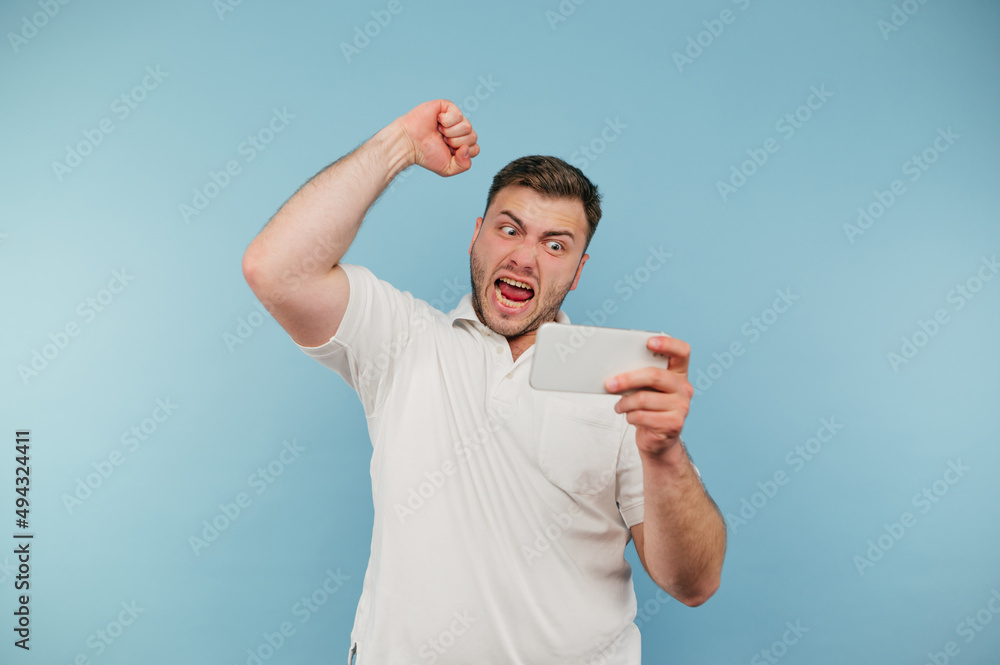 Angry man emotionally reacts to a loss with a smartphone in hand looks at the screen and screams in anger, isolated on a blue background Angry man emotionally reacts to a loss with a smartphone in hand looks at the screen and screams in anger, isolated on a blue background