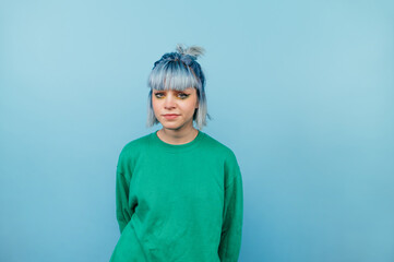 Portrait of unhappy girl in green sweatshirt and blue hair standing and looking at camera with serious face isolated on blue background.