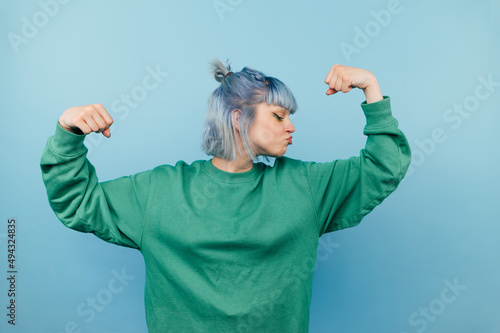 Funny hipster girl in green sweatshirt shows biceps and strength, kisses hand while standing on blue background,