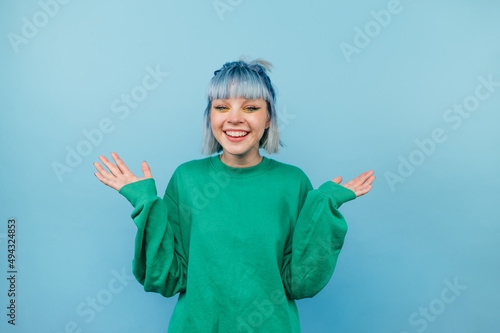 Positive teen girl with blue hair and in a green sweater rejoices with raised hands on a colored background.