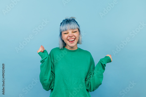 Joyful girl with blue hair in a green sweatshirt rejoices with a smile on his face and closed eyes on a blue background
