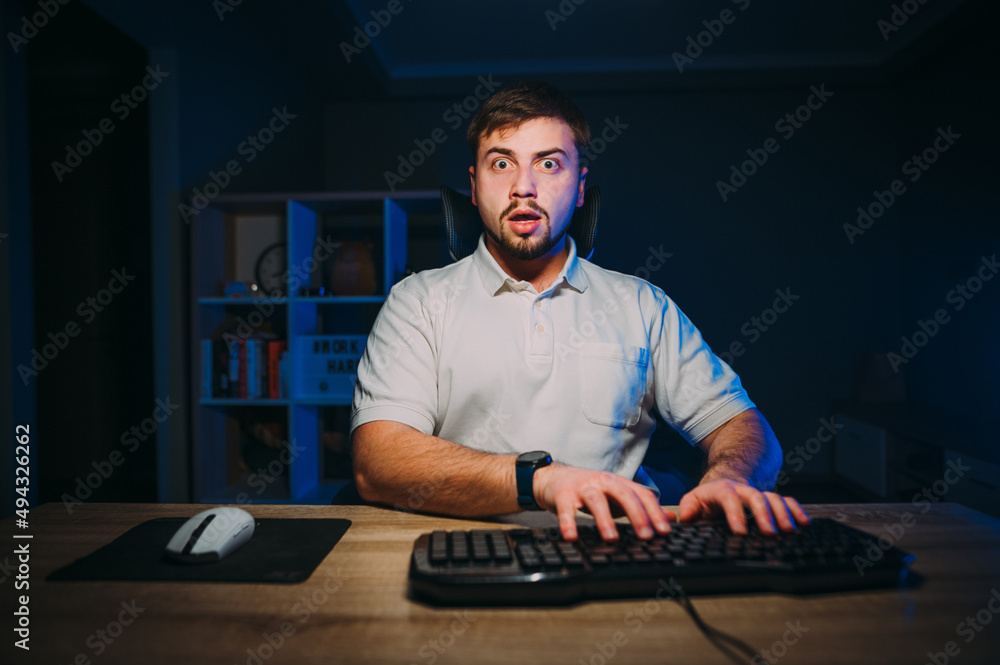 An emotional man with a beard in a T-shirt sits at the computer at night and works, looking at the camera with a shocked face. An emotional man with a beard in a T-shirt sits at the computer at night and works, looking at the camera with a shocked face.