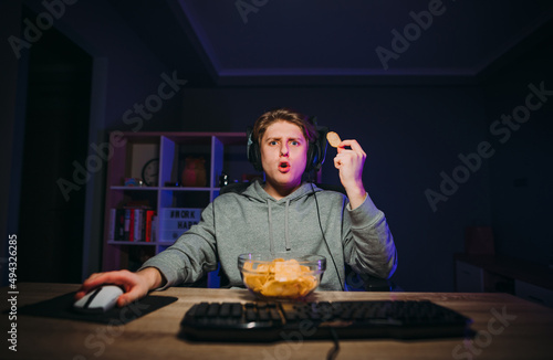 Shocked young man in a headset sits at night at the computer and eats chips from a plate with a surprised face staring at the screen.