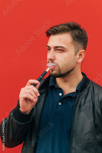 Handsome adult man with a beard smokes an IQOS on a background of a red wall and looks away.