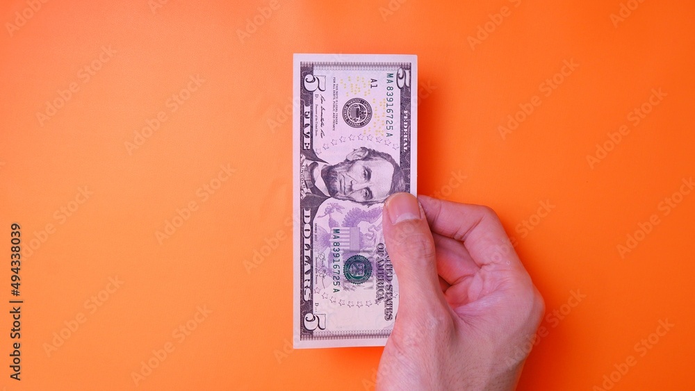 Man's hand is making a payment. Business Investment Economy Saving Loan Income Money and Finance concept. Male hand showing dollar cash on an orange background. Five Dollar 5 USD. Prosperity concept. Man's hand is making a payment. Business Investment Economy Saving Loan Income Money and Finance concept. Male hand showing dollar cash on an orange background. Five Dollar 5 USD. Prosperity concept.