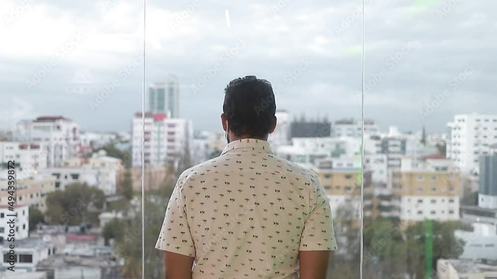 señor parada frente a la ventana con vista a la cuidad de espalda a la camara mientras nos acercamos, dolly in