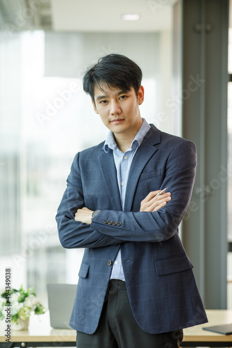 Portrait of young happy Asianbusinessman wearing grey suit and blue shirt standing in his office and smiling with arms crossed photo