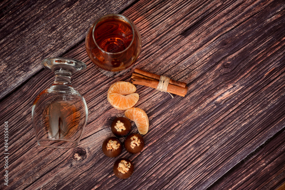 Two glasses of cognac, cinnamon, mandarin slices and chocolates on a wooden table.