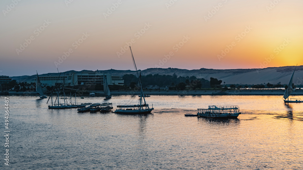 Evening Nile. Felucca and tourist boats on calm water. The sun hid behind a sand dune. The sky is highlighted in orange. Glare on the river. Egypt. Aswan