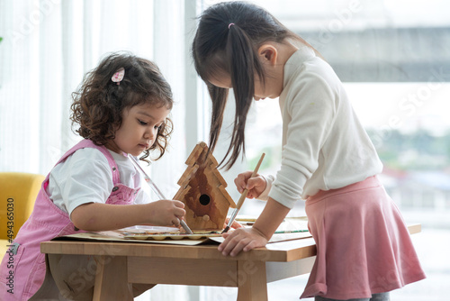Photo Two little girls help paint a wooden birdhouse in the room, education and learni