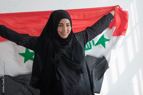 Happy Muslim woman wears black Hijab and Muslim traditional clothes holding irag flag, sunlight and shade on white wall, Muslim woman rights concept photo