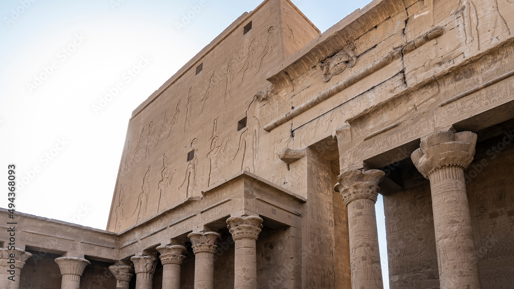 Fototapeta premium A fragment of the temple of Horus in Edfu. Columns with carved capitals, a wall with huge images of gods are visible. Blue sky. Egypt