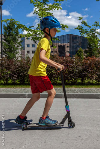 A young man in a yellow T-shirt and a blue helmet rides a stunt scooter through the city.