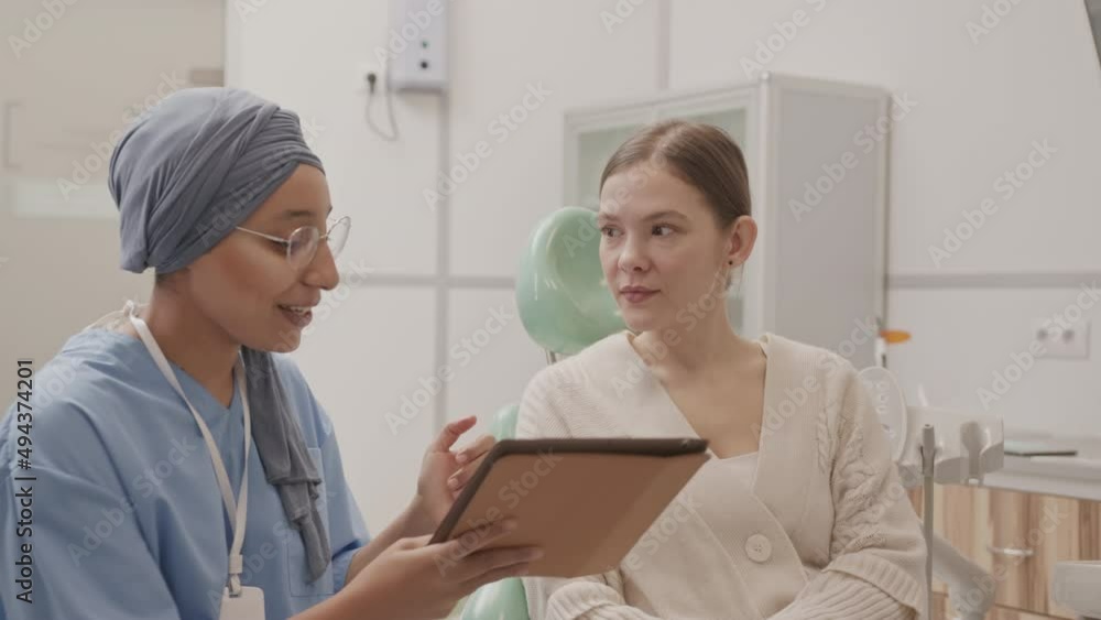 Medium slowmo of young Caucasian woman sitting at dental chair listening to female orthodontist in blue scrubs and headscarf holding tablet pc while talking about dental treatment Medium slowmo of young Caucasian woman sitting at dental chair listening to female orthodontist in blue scrubs and headscarf holding tablet pc while talking about dental treatment