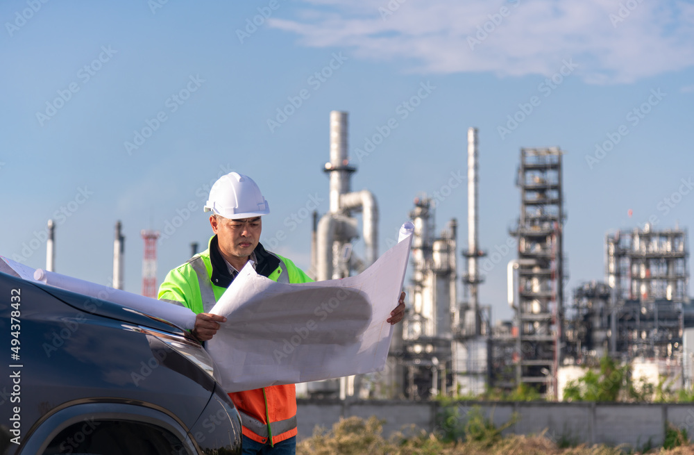 Engineer wearing uniform and helmet stand in front of the car hand holding blue print paper, inspection work plant site progress using radio communication to work orders with oil refinery background. Engineer wearing uniform and helmet stand in front of the car hand holding blue print paper, inspection work plant site progress using radio communication to work orders with oil refinery background.