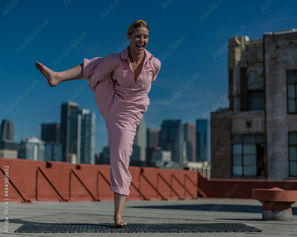 Young blonde woman in pink coveralls and barefoot practices yoga on a downtown Los Angeles Roof top with the LA skyline behind her Young blonde woman in pink coveralls and barefoot practices yoga on a downtown Los Angeles Roof top with the LA skyline behind her