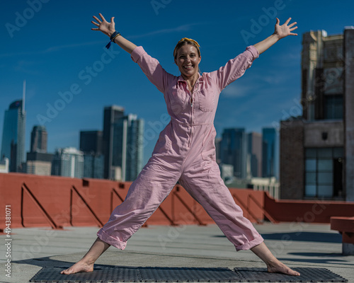 Young blonde woman in pink coveralls and barefoot practices yoga on a downtown Los Angeles Roof top with the LA skyline behind her