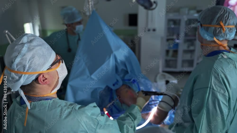 Chief surgeon holds and moves device input into patient's body. Medical team standing around the patient. Blurred backdrop. Chief surgeon holds and moves device input into patient's body. Medical team standing around the patient. Blurred backdrop.
