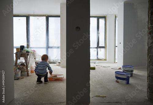 Side view of little adorable boy in colorful sweater doing repair at home with special brush for painting wall. Concept of preparing for drawing wall in white color in room at home.