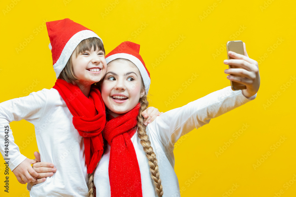 Fototapeta premium Youth Lifestyle Ideas. Couple of Caucasian Girlfriends Taking Selfie While Wearing New Year Christmas Santa Hats And Posing Against Seamless Yellow Background.