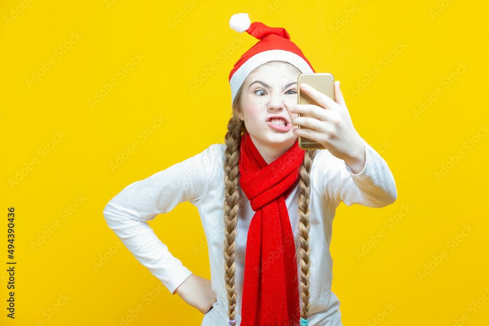 Fototapeta premium Funny Winsome Caucasian Teenage Girl With Cellphone Taking Selfie While Making Gimmick And Wearing Christmas Santa Hat And Posing Against Yellow Background.
