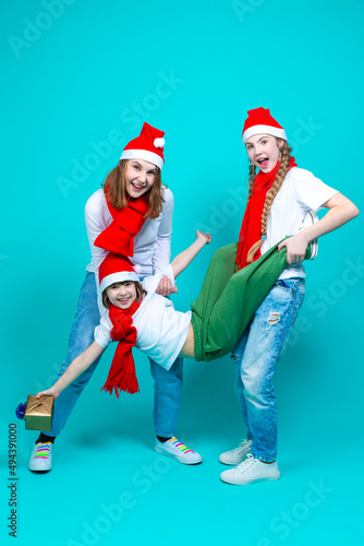 Three Positive Caucasian Girls With Santa Hats Having Fun While Lifting Teenage Girl Holding Wrapped Gift Box Against Trendy Green-Blue Background.