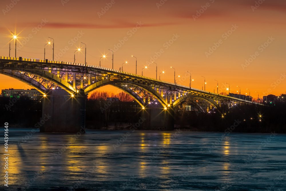 Naklejka premium Bridge at sunset, with yellow illumination, frozen river, ice.