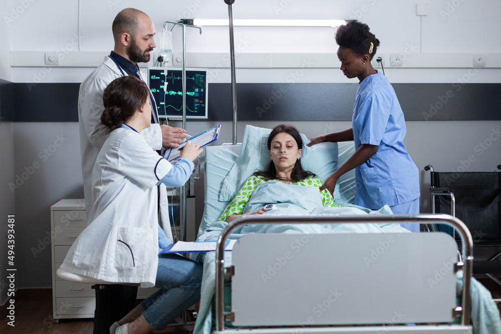 Team of doctors discussing patient medical history on clipboard before surgery while nurse is giving medical care. Woman with low spo2 saturation connected to iv drip line and monitor showing vitals. Team of doctors discussing patient medical history on clipboard before surgery while nurse is giving medical care. Woman with low spo2 saturation connected to iv drip line and monitor showing vitals.
