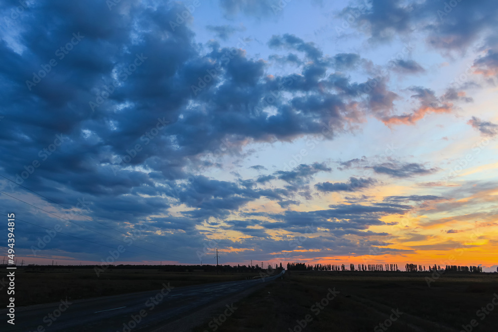 A beautiful bright sunset in the countryside. The orange-blue sky looms over the country road and fields.