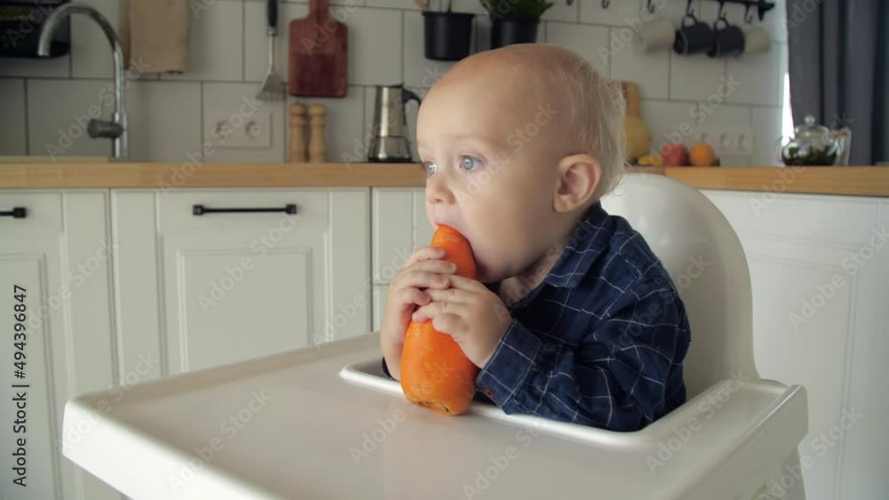 Cute baby eating carrot in a white kitchen. Healthy nutrition for kids. Bio carrot as first solid food for infant. Little boy biting raw vegetable. Cute baby eating carrot in a white kitchen. Healthy nutrition for kids. Bio carrot as first solid food for infant. Little boy biting raw vegetable.