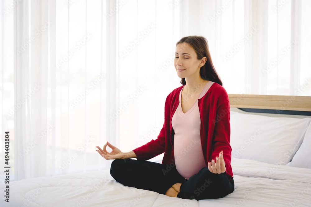 Pregnant woman sitting on bed and practice yoga in modern bedroom.