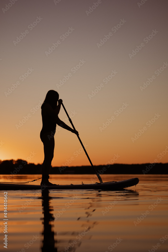 Low angle of woman silhouette standing on paddle board with paddle in hands on lake at sunset time with trees in background in summer. Active lifestyle.